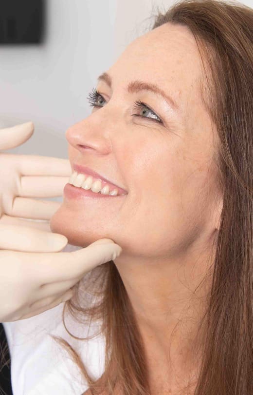 Close-up of a smiling woman looking upward while a medical professional wearing gloves gently examines her chin during a cosmetic consultation.